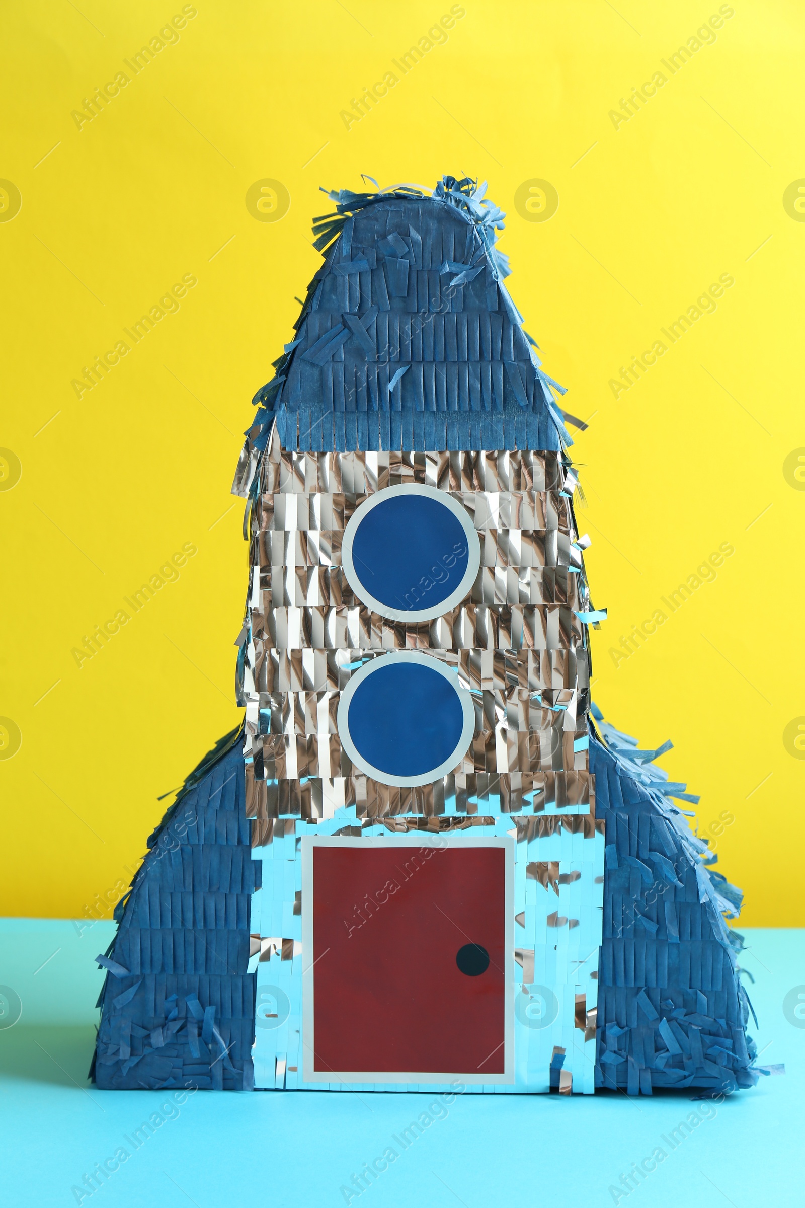 Photo of Beautiful pinata in shape of rocket on blue table against yellow background