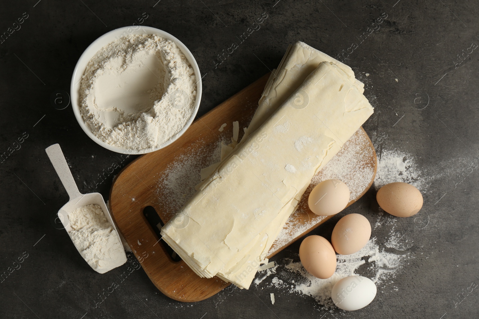 Photo of Raw puff pastry dough, eggs and flour on dark textured table, flat lay