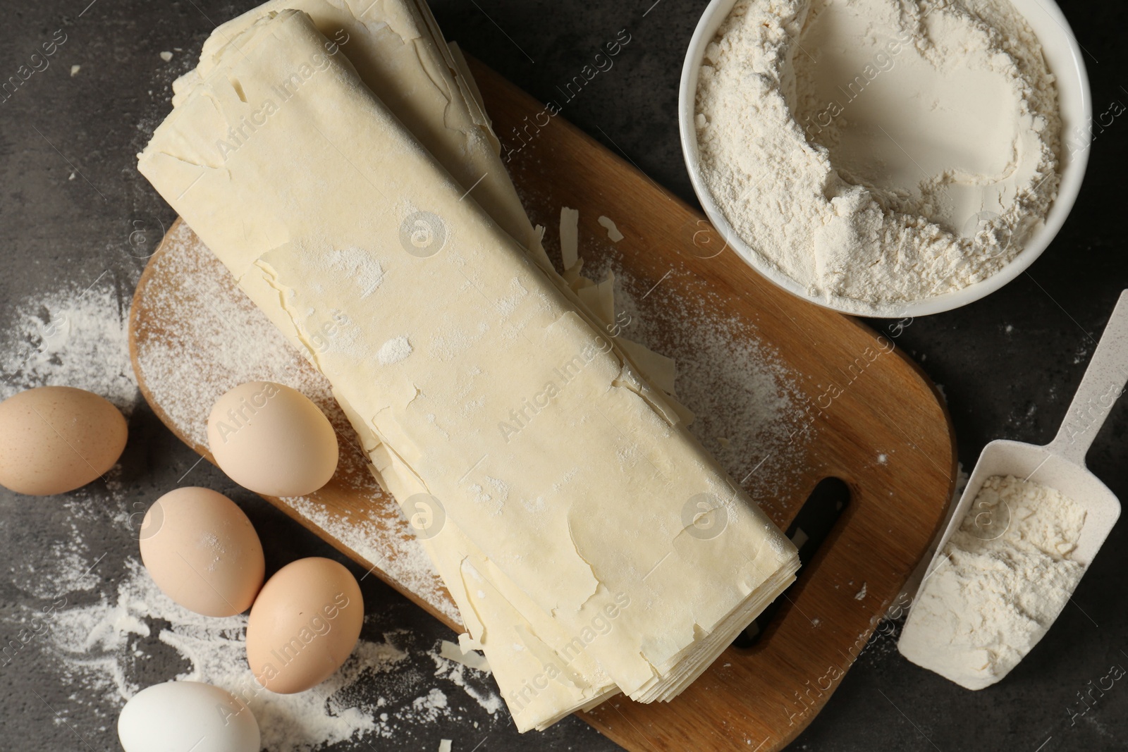 Photo of Raw puff pastry dough, eggs and flour on dark textured table, flat lay