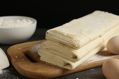 Photo of Board with raw puff pastry dough, eggs and flour on dark textured table, closeup