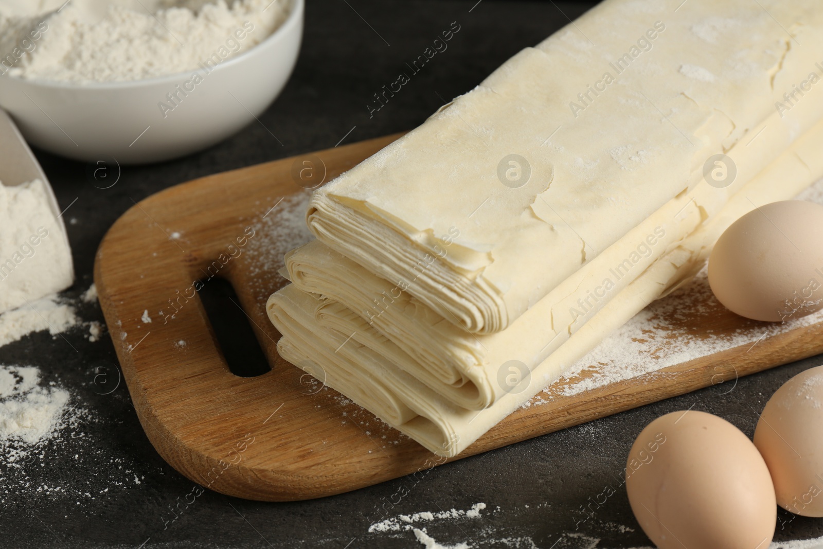 Photo of Board with raw puff pastry dough, eggs and flour on dark textured table, closeup