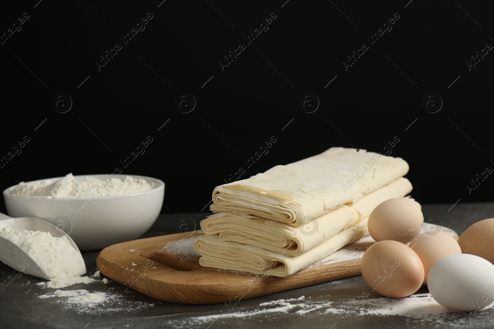 Photo of Board with raw puff pastry dough, eggs and flour on dark textured table against black background