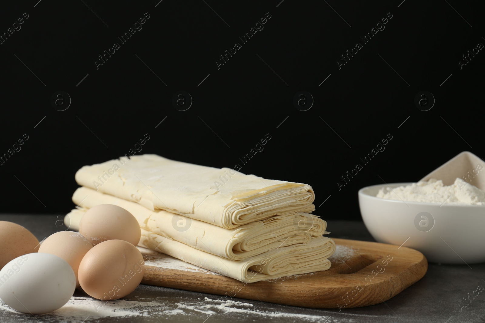 Photo of Board with raw puff pastry dough, eggs and flour on dark textured table against black background