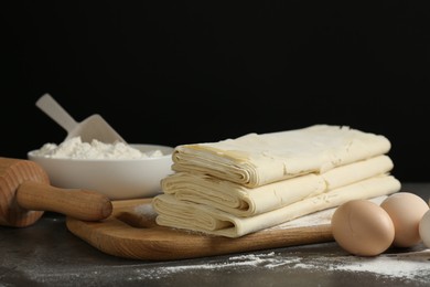 Photo of Board with raw puff pastry dough, eggs and flour on dark table against black background