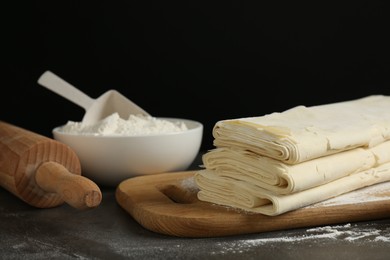 Photo of Board with raw puff pastry dough, rolling pin and flour on dark table against black background, closeup