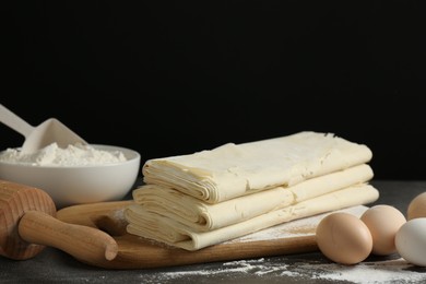 Photo of Board with raw puff pastry dough, eggs and flour on dark table against black background