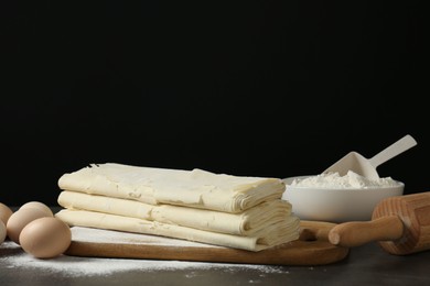 Photo of Board with raw puff pastry dough, eggs and flour on dark table against black background