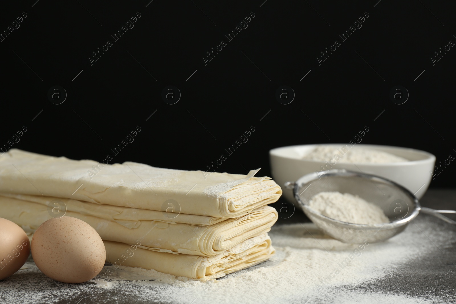 Photo of Stack of raw puff pastry dough, eggs and flour on dark table against black background, closeup