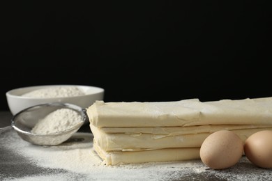 Stack of raw puff pastry dough, eggs and flour on dark table against black background, closeup