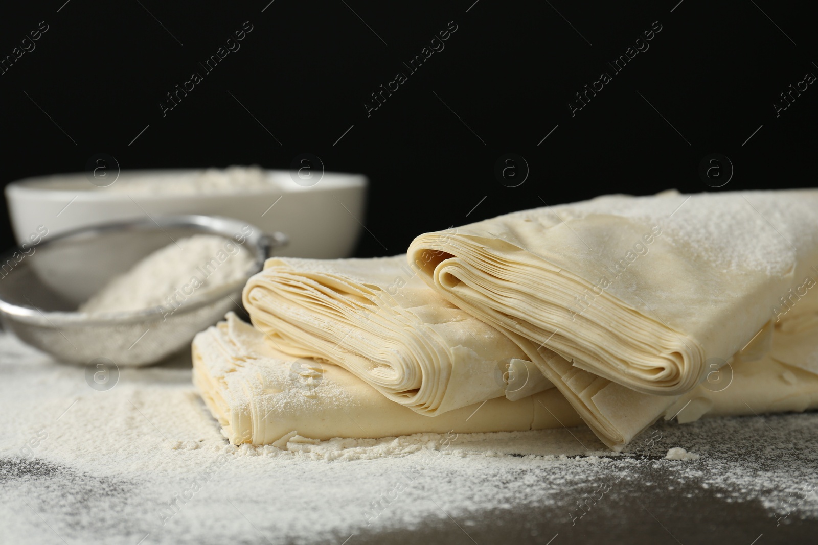 Photo of Raw puff pastry dough and flour on dark table against black background, closeup