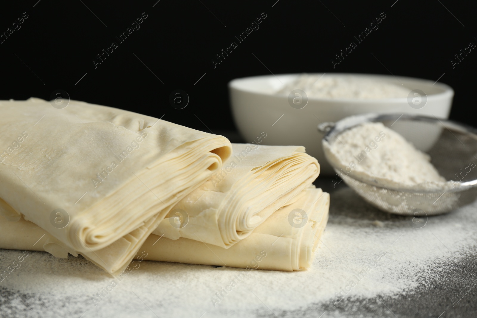 Photo of Raw puff pastry dough and flour on dark table against black background, closeup