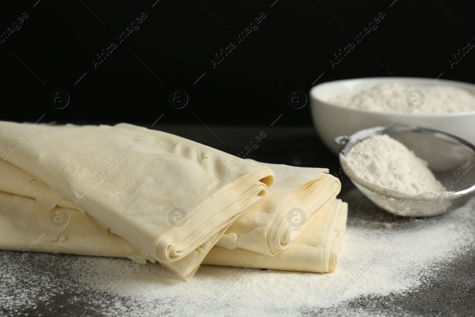 Photo of Raw puff pastry dough and flour on dark table against black background, closeup