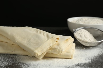 Photo of Raw puff pastry dough and flour on dark table against black background, closeup