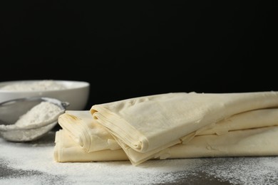 Photo of Raw puff pastry dough and flour on dark table against black background, closeup