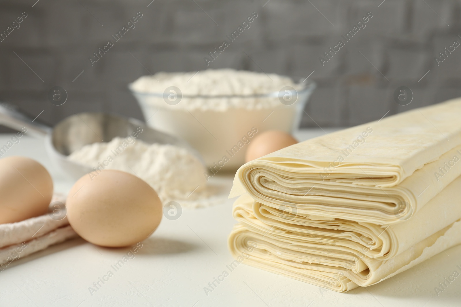 Photo of Stack of raw puff pastry dough, eggs and flour on white table, closeup