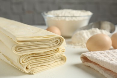 Photo of Stack of raw puff pastry dough, eggs and flour on white table, closeup