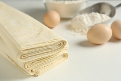 Photo of Stack of raw puff pastry dough, eggs and flour on white table, closeup