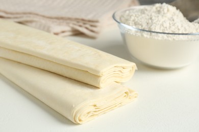 Photo of Raw puff pastry dough and flour on white table, closeup