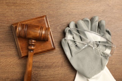 Photo of Accident at work concept. Gavel, protective gloves and goggles on wooden table, flat lay