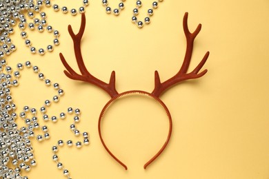 Photo of Beautiful reindeer headband and Christmas beads on pale yellow background, top view