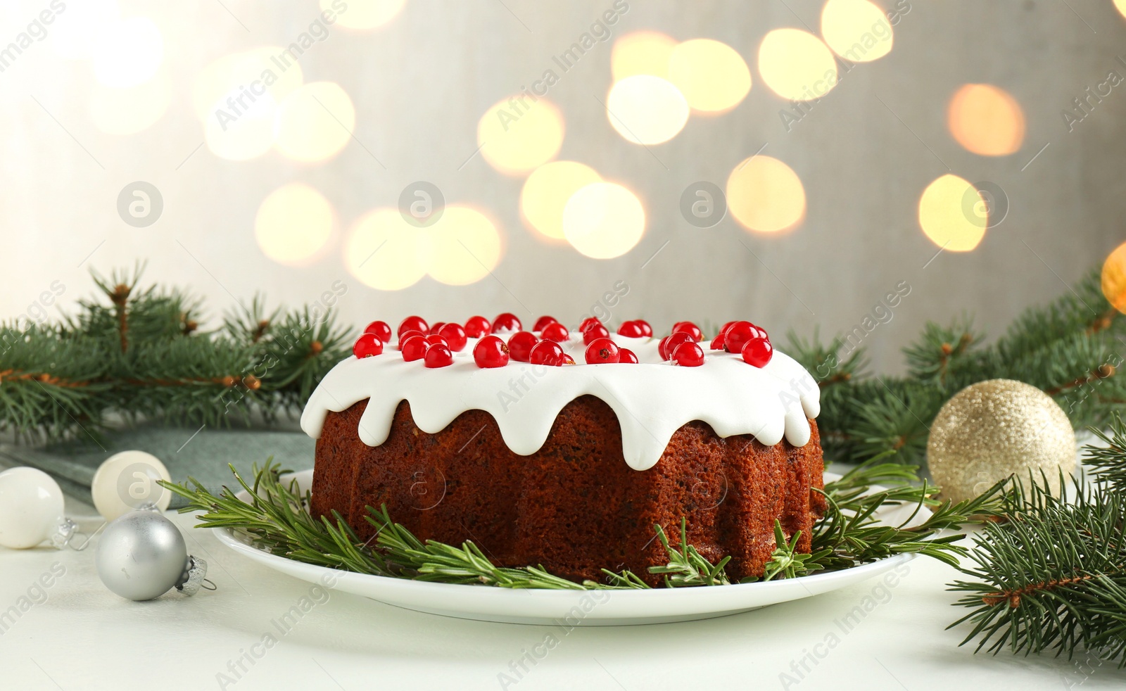 Photo of Beautifully decorated Christmas cake, fir branches and baubles on white table against blurred lights