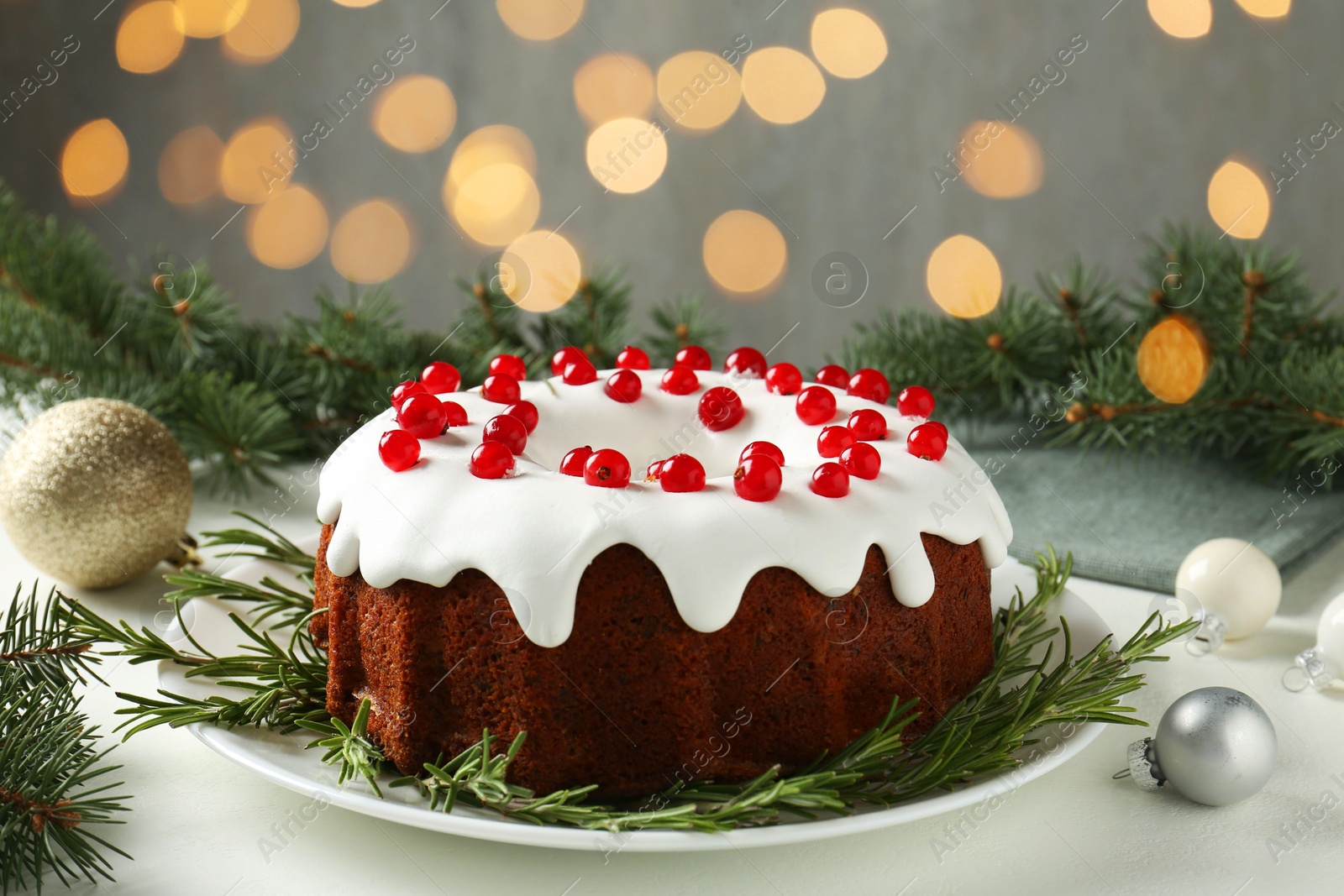 Photo of Beautifully decorated Christmas cake, fir branches and baubles on white table against blurred lights