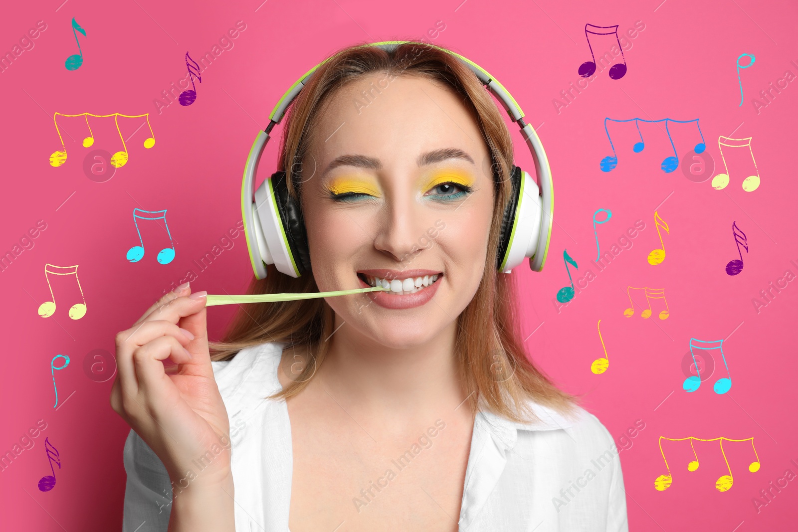 Image of Fashionable young woman with bright makeup and headphones chewing bubblegum on pink background. Music notes around her