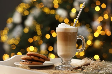 Photo of Aromatic coffee in glass, tasty cookies and cinnamon sticks on tray against blurred Christmas lights, bokeh effect