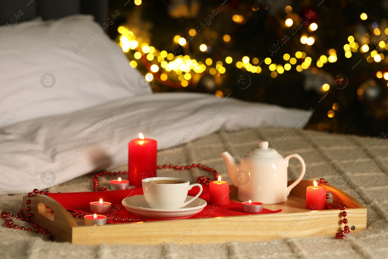 Photo of Aromatic tea in cup, teapot and burning candles on bed against blurred Christmas lights, bokeh effect