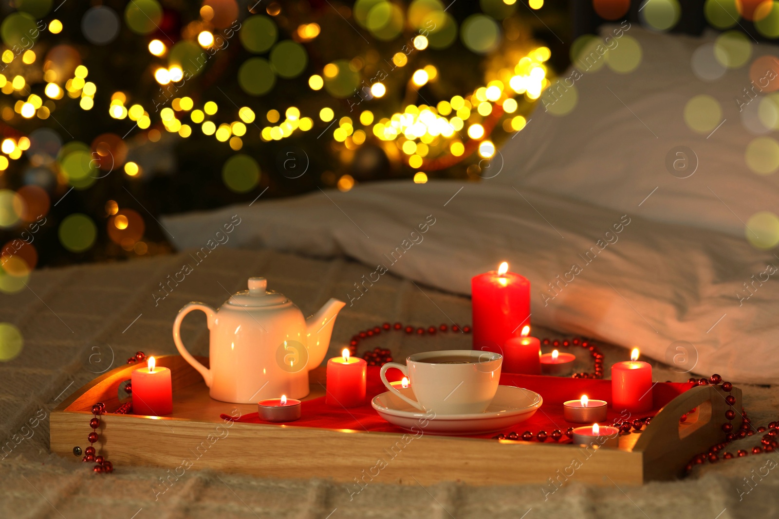 Photo of Aromatic tea in cup, teapot and burning candles on bed against blurred Christmas lights, bokeh effect