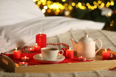 Photo of Aromatic tea in cup, teapot and burning candles on bed against blurred Christmas lights, bokeh effect