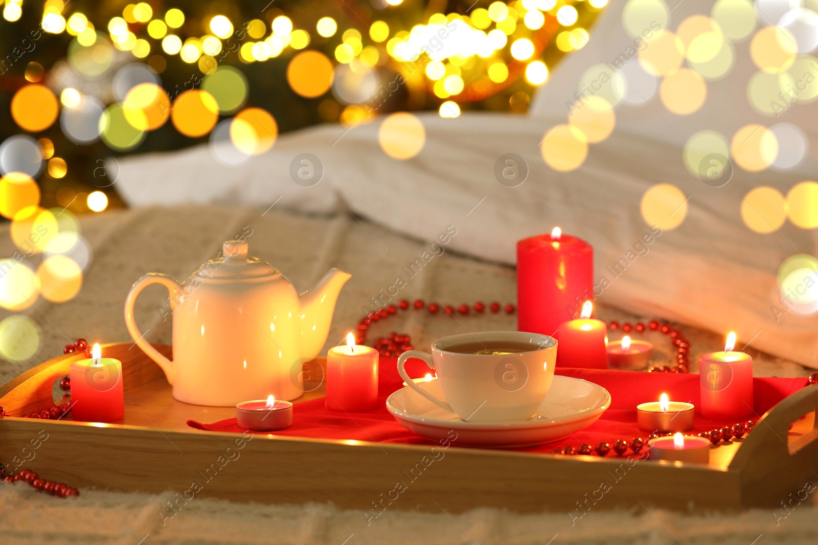 Photo of Aromatic tea in cup, teapot and burning candles on bed against blurred Christmas lights, bokeh effect