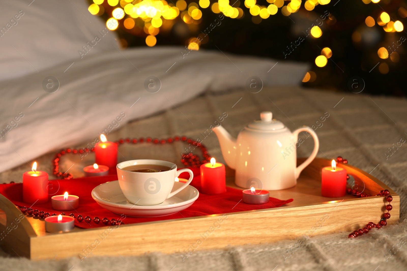 Photo of Aromatic tea in cup, teapot and burning candles on bed against blurred Christmas lights, bokeh effect