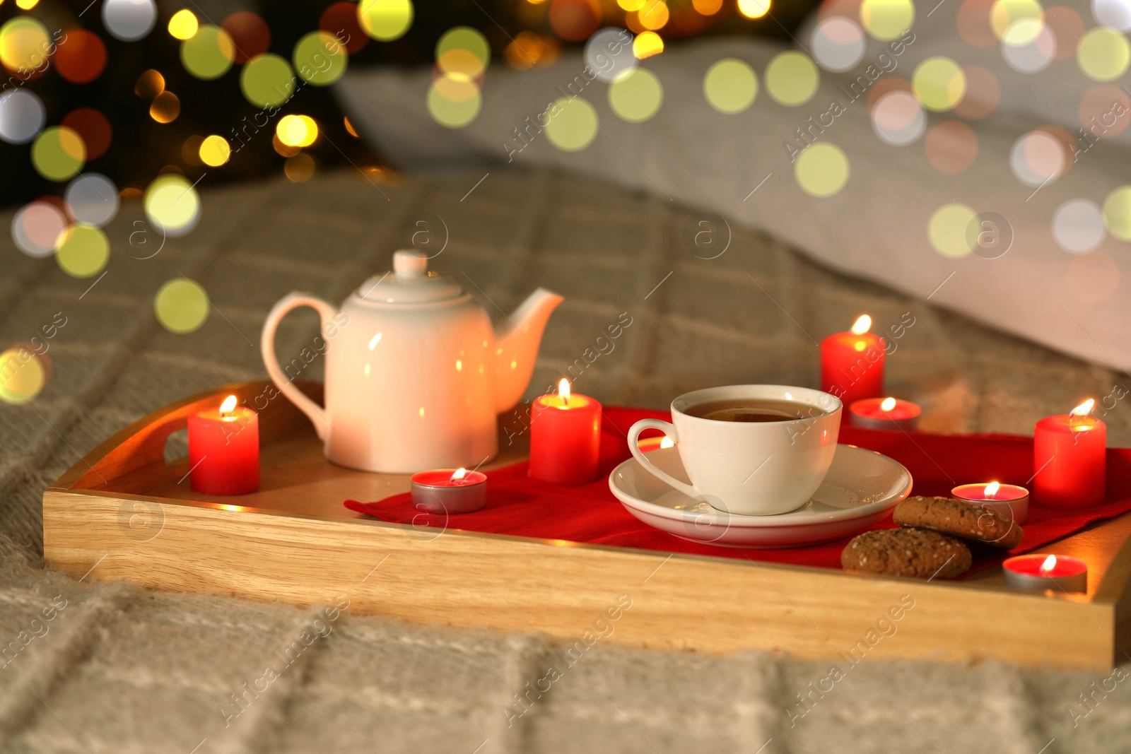 Photo of Aromatic tea in cup, cookies, teapot and burning candles on bed against blurred Christmas lights, bokeh effect