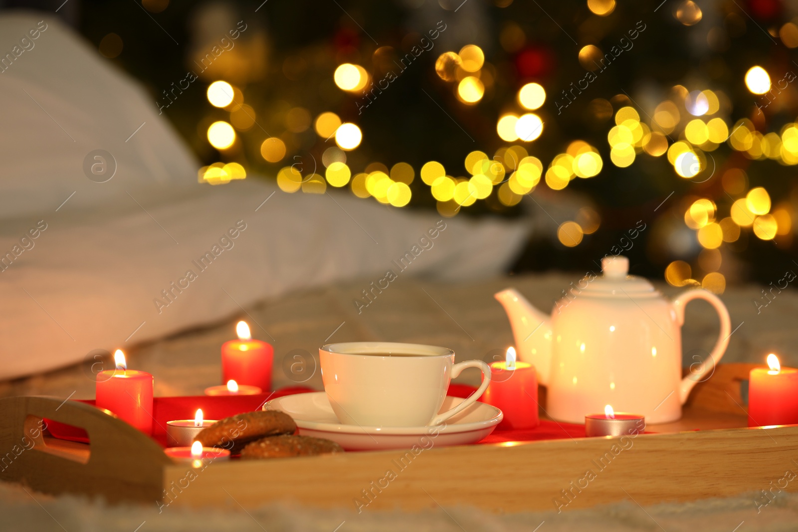 Photo of Aromatic tea in cup, cookies, teapot and burning candles on bed against blurred Christmas lights, bokeh effect