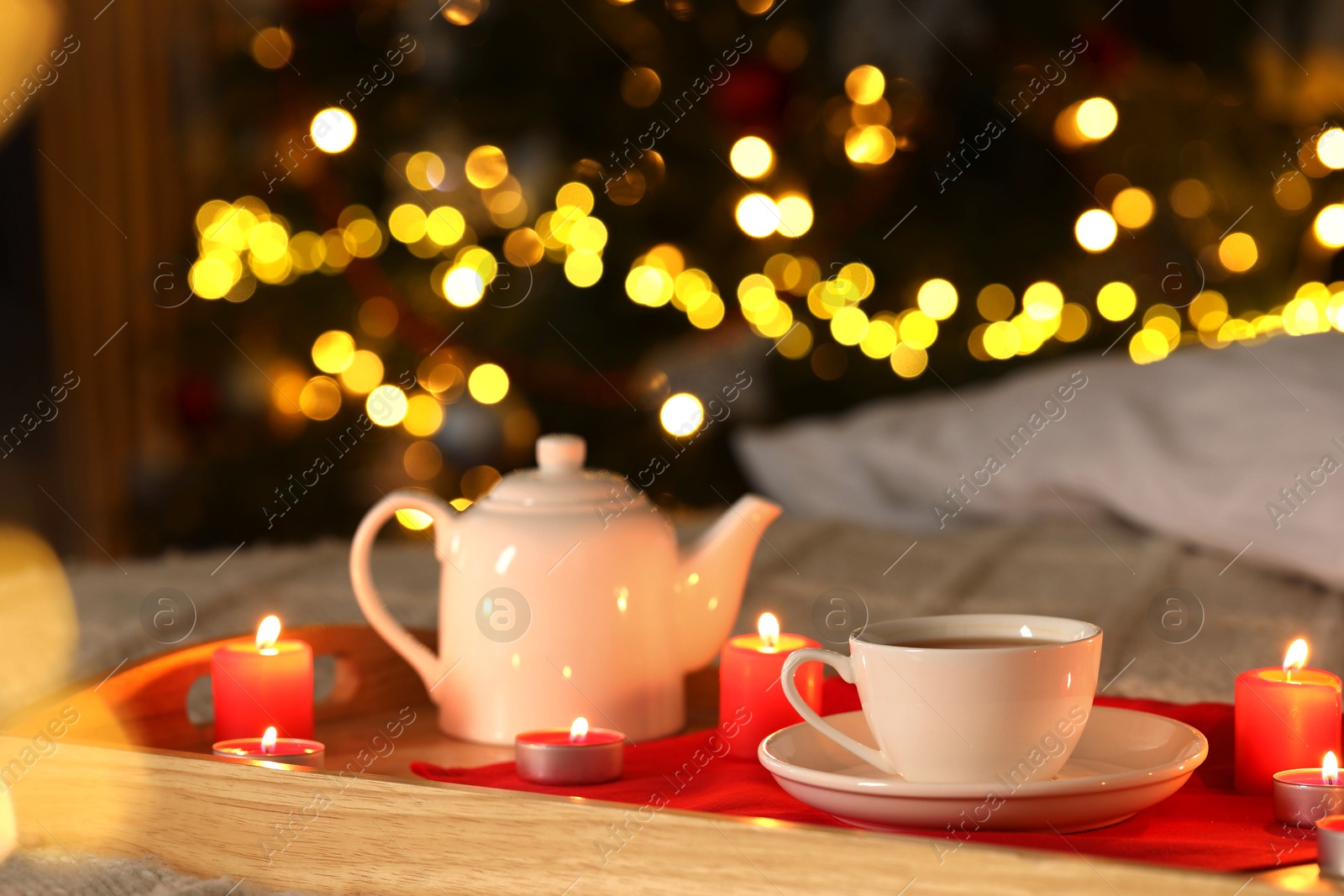 Photo of Aromatic tea in cup, teapot and burning candles on bed against blurred Christmas lights, closeup. Bokeh effect