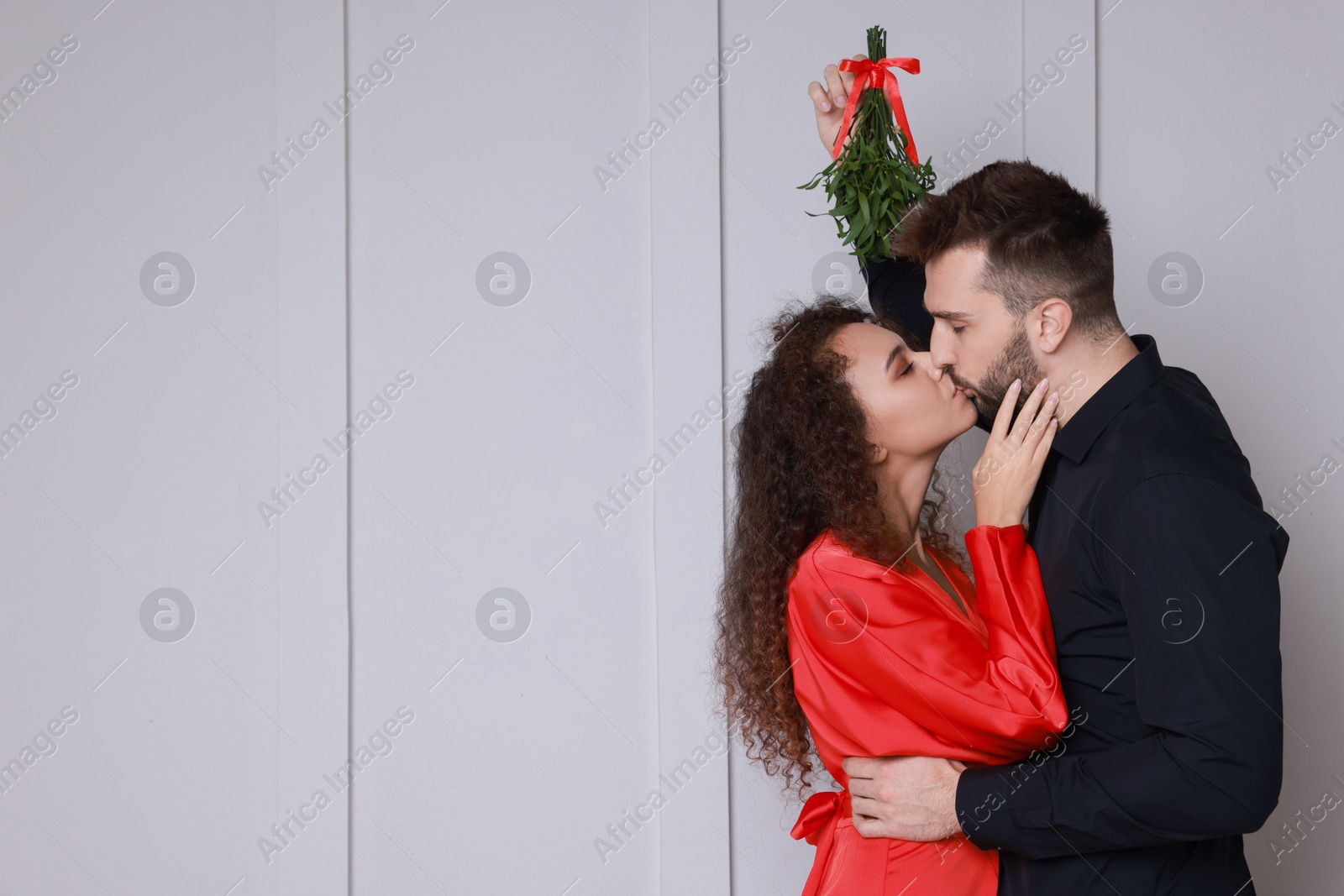 Photo of Happy couple kissing under mistletoe bunch near light grey wall. Space for text