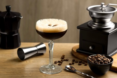 Photo of Delicious espresso martini in glass, coffee beans, manual grinder and jigger on wooden table, closeup