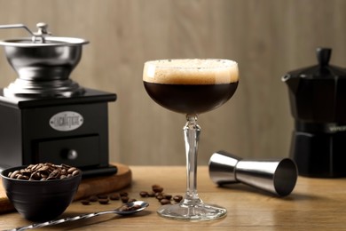 Photo of Delicious espresso martini in glass, coffee beans, manual grinder and jigger on wooden table, closeup