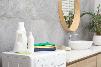 Photo of Laundry detergents, brush and stacked clean clothes on washing machine in bathroom