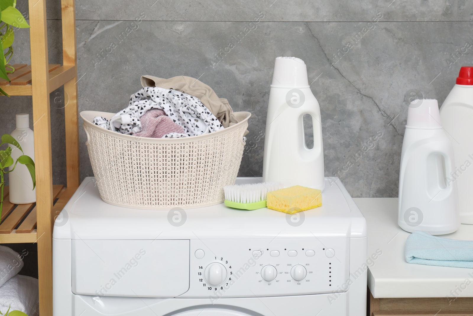 Photo of Detergents, basket with laundry, brush, sponge and washing machine indoors