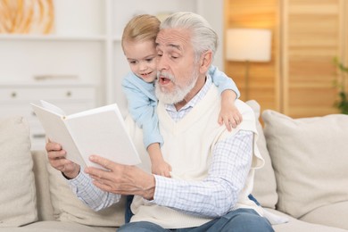 Photo of Grandpa and his granddaughter reading book together on sofa at home