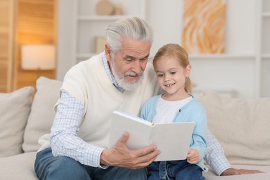 Photo of Grandpa and his granddaughter reading book together on sofa at home