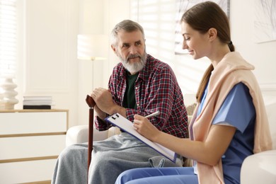 Photo of Caregiver examining senior man on sofa indoors. Home health care service