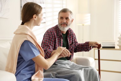 Photo of Caregiver supporting senior man on sofa indoors. Home health care service