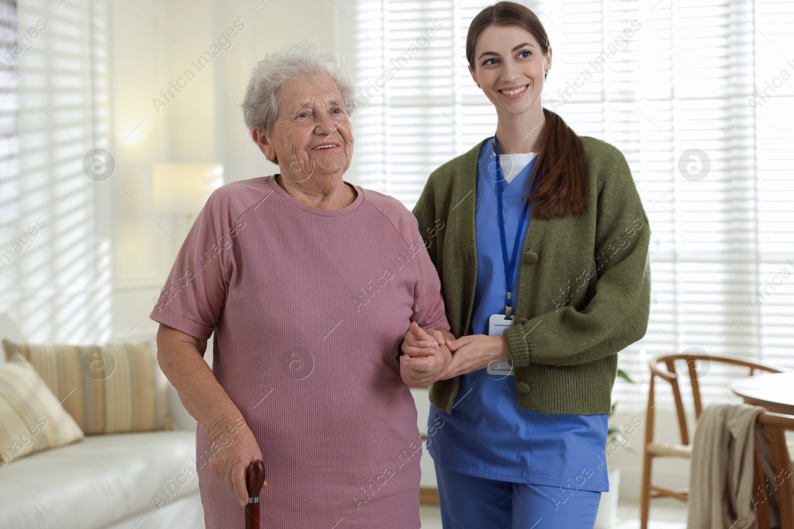 Photo of Caregiver assisting senior woman with walking cane indoors. Home health care service