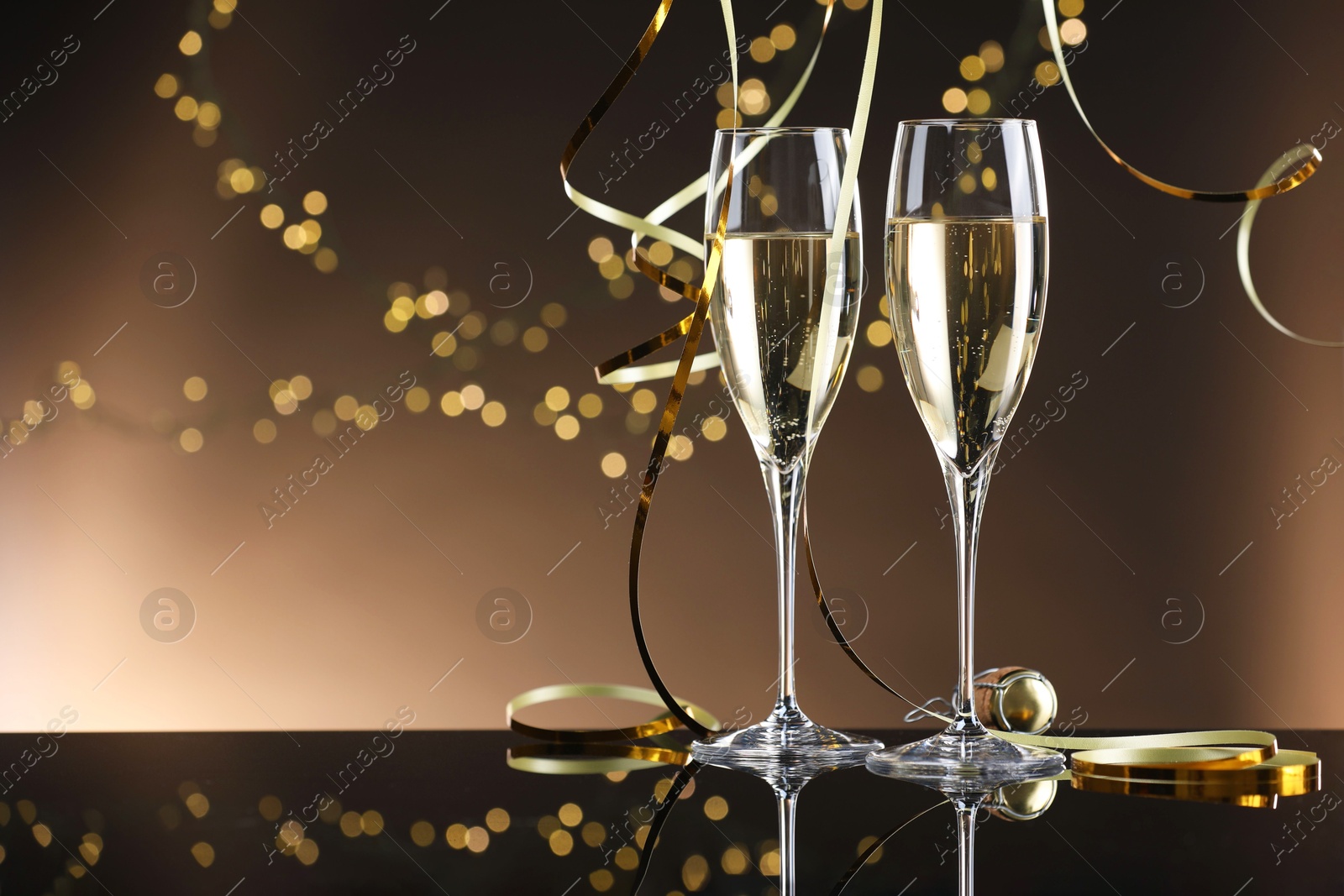 Photo of Champagne in glasses and party streamers on mirror surface against brown background with blurred lights, space for text