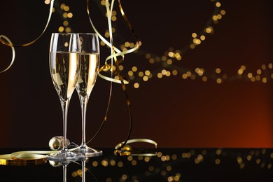 Photo of Champagne in glasses and party streamers on mirror surface against color background with blurred lights, space for text