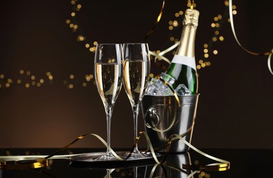 Photo of Champagne in glasses and ice bucket with bottle on mirror surface against brown background with blurred lights