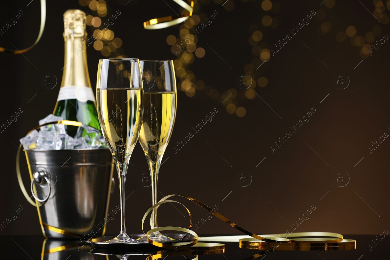 Photo of Champagne and bucket of ice on mirror surface against brown background with blurred lights, space for text